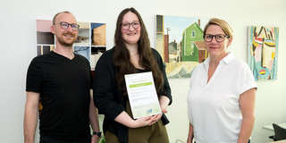 Drei Personen stehen gemeinsam in einem Büro; die mittlere Person hält ein Zertifikat in den Händen.