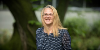 The portrait photo shows Prof. Angela Madeo. She has blonde, medium-length hair and is wearing a blue and white top. Bushes and tree trunks are blurred in the background.