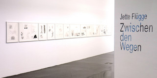 Das Foto zeigt einen Ausstellungsraum mit mehreren Bilderrahmen auf einer weißen Wand. Rechts an einer Wand steht in blauer Schrift: "Jette Flügge. Zwischen den Wegen."