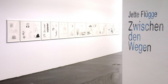 Das Foto zeigt einen Ausstellungsraum mit mehreren Bilderrahmen auf einer weißen Wand. Rechts an einer Wand steht in blauer Schrift: "Jette Flügge. Zwischen den Wegen."