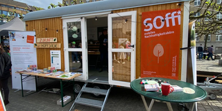 Abgebildet ist das mobile Nachhaltigkeitsbüro "Soffi", ein Container mit einer geöffneten Glastür, Holzverkleidung und einem Banner des Nachhaltigkeitsbüros. Vor dem Container steht ein Tisch mit Infomaterialien.