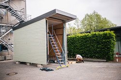 Eine Person arbeitet neben einem kleinen, unfertigen Gebäude mit grünen Wänden und einem flachen Dach. Das Gebäude steht in einem Hinterhof. Auf dem Boden liegen Werkzeuge und Baumaterialien.