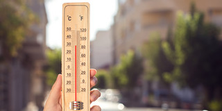 Eine Hand hält ein Thermometer, das etwa 30 Grad Celsius anzeigt. Im Hintergrund verschwommen eine Stadt im Sommer.