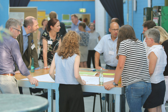 The picture shows a group of people standing around a table and discussing.