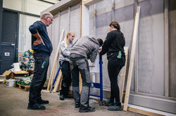 Zwei Frauen halten ein Gerüst, ein Mann steht davor und scheint an dem Gerüst zu schrauben. Ein weiterer Mann steht daneben und schaut zu. Die Personen sind umgeben von Werkzeug und Baumaterialien.