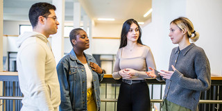 Das Bild zeigt vier Studierende, die in einem Halbkreis zueinander stehen. Drei Personen der Studierenden richten alle den Blick auf eine sprechende Person.