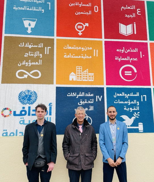 Drei Studierende stehen vor einem Plakat der Universität Mossul.
