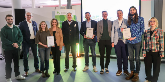 Foto: Mehrere Personen stehen gemeinsam für ein Gruppenfoto mit Urkunden vor einer Leinwand und einem Banner der TU Dortmund.