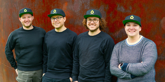 Maximilian Spiekermann, Jens Ehlhardt, Max Krause und Marén Schwandt (v.l.n.r.) stehen vor einer Rostroten Wand. Alle vier Personen tragen eine rote Cappy mit einem gelben Tropfensymbol und der Unterschrift "Simplyfined".