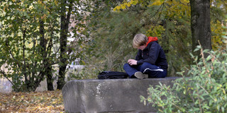 A boy sits alone cross-legged on a flat stone. His bag lies in front of him, his head is lowered so that his face is unrecognizable.