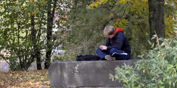 A boy sits alone cross-legged on a flat stone. His bag lies in front of him, his head is lowered so that his face is unrecognizable.