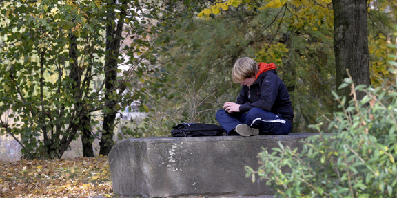 A boy sits alone cross-legged on a flat stone. His bag lies in front of him, his head is lowered so that his face is unrecognizable.