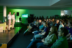 Foto: Zwei Personen stehen auf einer Bühne und sprechen vor einem zuhörenden Publikum.