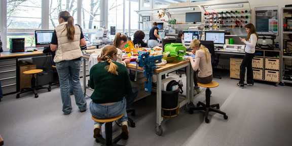 Foto: Mehrere Personen arbeiten gemeinsam in einer Werkstatt mit 3D-Druckern, Computern und verschiedenen Materialien.