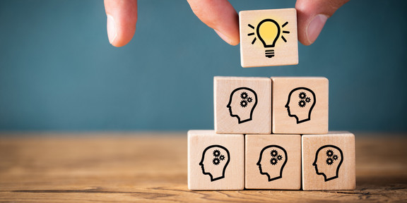 A hand placing a wooden cube with the symbol of a glowing light bulb as the last top tile on a pyramid of other wooden cubes, all of which show the same motif: The schematic representation of a head, each with three cogwheels inside.