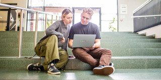 Zwei junge Erwachsene sitzen auf der Treppe im Foyer des Seminarraumgebäudes der TU Dortmund und schauen auf einen grauen Laptop.