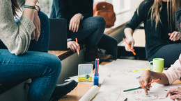 Vier Menschen sitzen an einem Tisch, die Gesichter sind nicht im Bild. Sie halten Stifte in der Hand und sehen nachdenkend aus. Eine Hand schreibt etwas auf das Plakat auf dem Tisch, welches sehr unstrukturiert gestaltet ist. Auf dem Tisch sind noch mehr Stifte und eine Tasse. Auf der Sitzbank erkennt man einen geöffneten Laptop und einen Rucksack im Hintergrund.