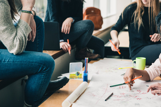 Four people are sitting at a desk writing something on a poster. There are also pencils and a cup on the table.