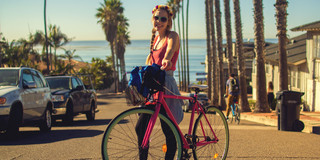 Junge Frau schiebt ein Fahrrad über eine Straße, im Hintergrund Palmen