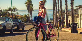 Young woman pushing a bicycle across a street, palm trees in the background