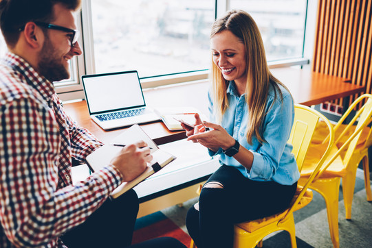 a man and a woman are sitting at a desk facing each other. On the desk is an open laptop