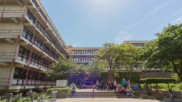 3: Chemie und Chemische Biologie Chemiegebäude: Ein großes, breites, graues Gebäude mit Fensterfronten und Balkonen vor den Fenstern, die mit roten Metallstangen abgegrenzt werden. In der Mitte eine Treppe, auf der Menschen sitzen und links vor der Treppe Fahrräder in Fahrradständern.