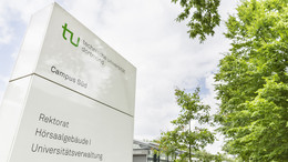 A white sign with TU Dortmund logo stands next to green trees.