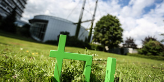 Im Vordergrund steht das TU-Logo im Gras, im Hintergrund das Audimax.