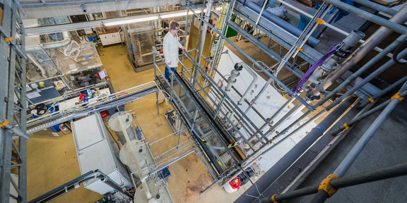 Laboratory_02 A person with a white coat stands in a laboratory with several large containers and metal pipes.