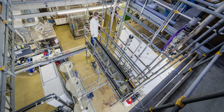 Eine Person mit weißem Kittel steht in einem Labor mit mehreren großen Behältern und Metallrohren.