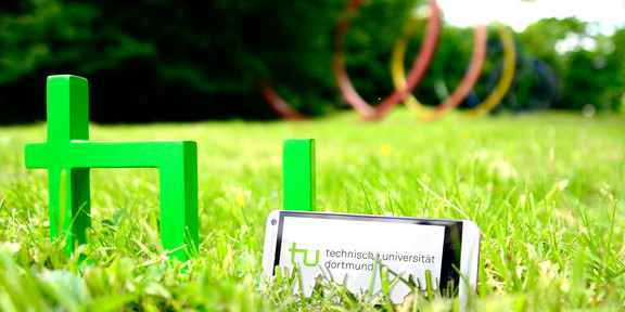 A smartphone standing in the grass next to the TU logo, the spectral rings can be seen in the background.