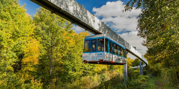 H-Bahn_04 Die H-Bahn fährt an einem Weg entlang mit grünen Bäumen.