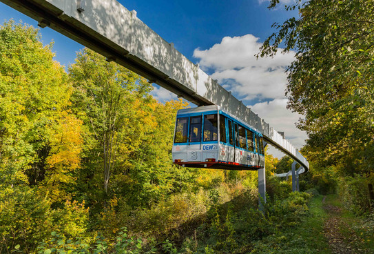 H-Bahn_04 Die H-Bahn fährt an einem Weg entlang mit grünen Bäumen.