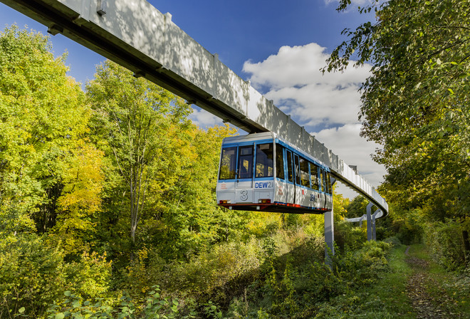 The H-Bahn runs along a path with green trees.