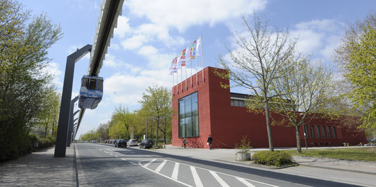 Rotes Gebäude umgeben von Bäumen mit blauem Himmel und wenigen Wolken. Links im Bild die H-Bahn.