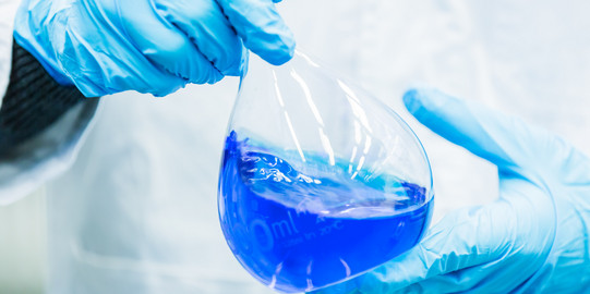 Detailaufnahme Messkolben Zwei Hände in blauen Gummihandschuhen halten einen Messkolben, der mit blauer Flüssigkeit gefüllt ist.