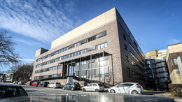 2: Physik Neues Physikgebäude: Ein modernes grau-braun verklinkertes Gebäude mit flachem Dach, vier Stockwerken und einem Vordach. Davor eine Straße, an der Autos hintereinander parken.