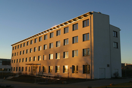 E-Technik-Gebäude Gebäude E-Technik: Ein hohes weißes Gebäude, das von der Sonne angestrahlt wird. Vor dem Gebäude eine Rampe und über dem Eingang ein Vordach.